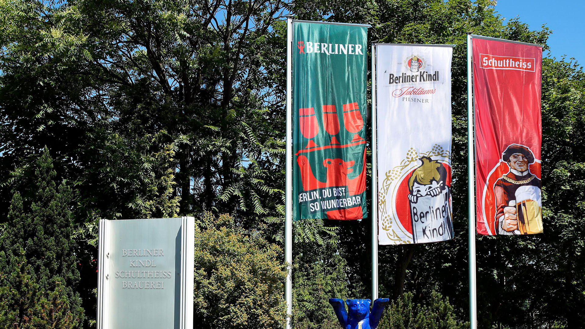 Drei vertikale Banner flattern vor einem bewaldeten Hintergrund im Wind.
