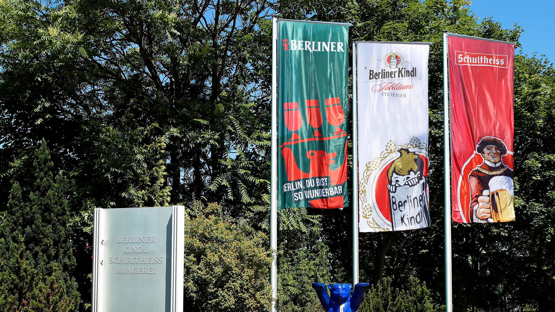 Drei vertikale Banner flattern vor einem bewaldeten Hintergrund im Wind.