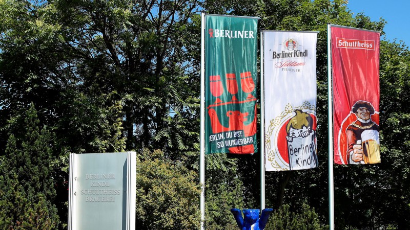 Ein Glas gefüllt mit goldgelbem Bier trägt ein Etikett mit einer lächelnden Figur in historischer Kleidung, die ein Bierkrug hält.
