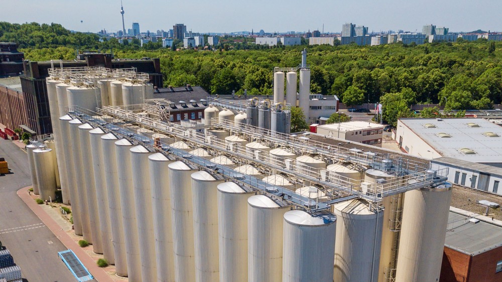 Ein Luftbild zeigt eine Anlage mit vielen großen grauen Silos, umgeben von Bäumen in einer städtischen Umgebung.