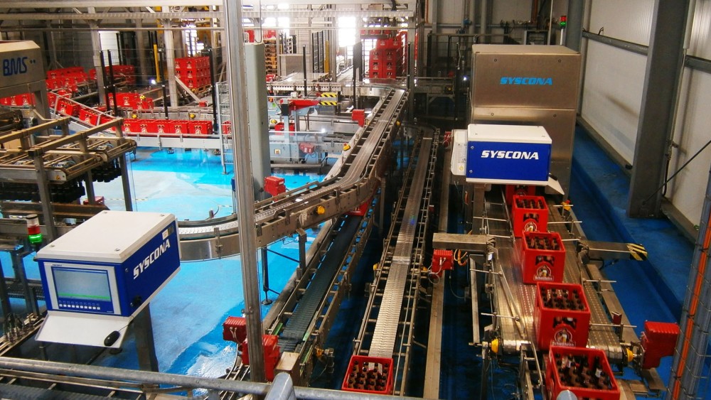 Eine Flaschenabfüllanlage in einer Fabrikhalle mit vielen Förderbändern und Maschinen. Leere und vollständig gefüllte Kisten bewegen sich durch die Anlage.