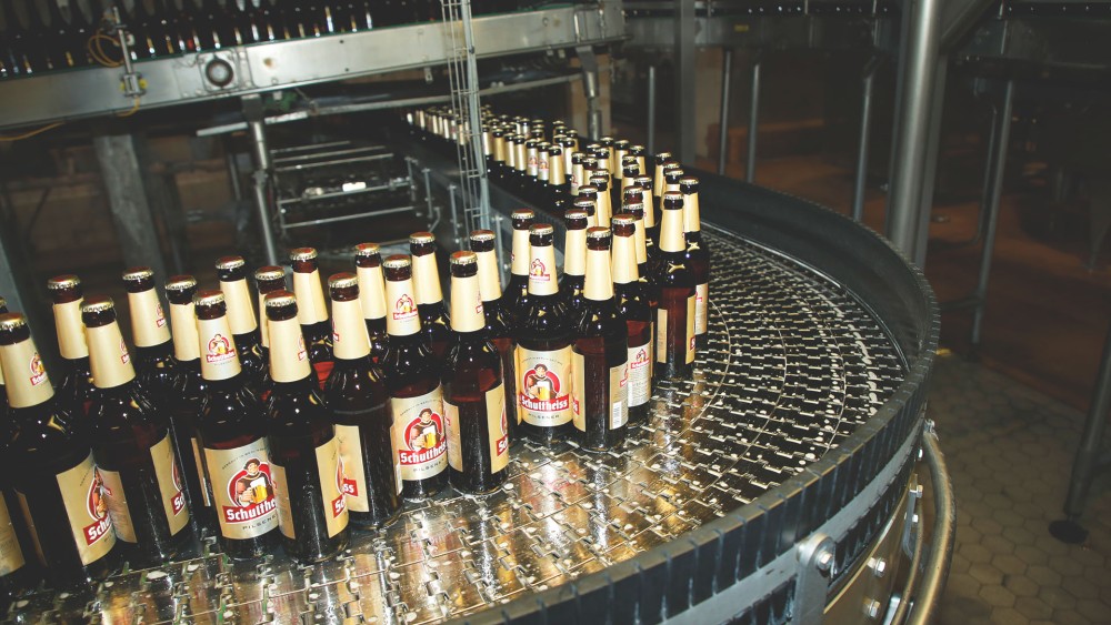 Bierflaschen bewegen sich auf einem Fließband in einer Brauerei. Sie sind dicht aneinandergereiht.