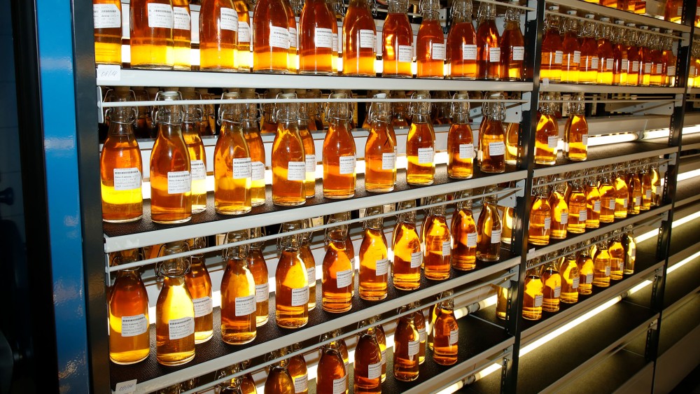 Reihen von Glasflaschen mit orangefarbenem Inhalt sind auf Metallregalen aufgestellt. Jede Flasche trägt ein weißes Etikett.