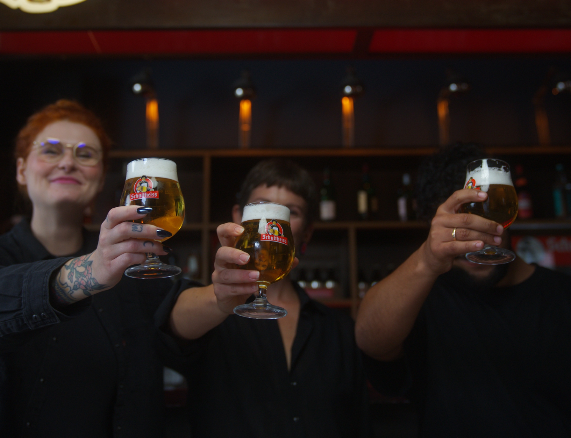 Drei Personen halten ein Schulheiss Bierglas in die Luft, dahinter ein Regal mit flaschen und Gläsern.